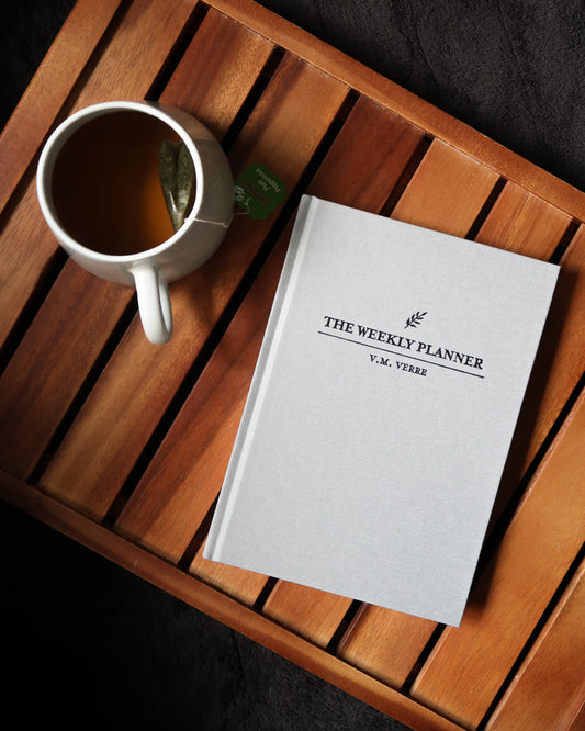 A light grey copy of The Weekly Planner by V.M. Verre next to a cup of tea in a beautiful mug.