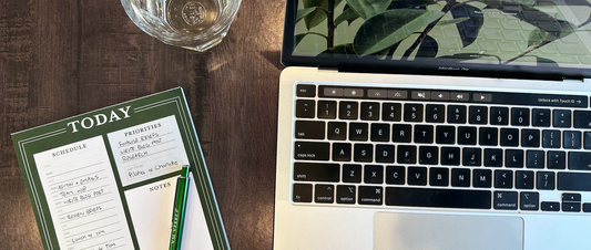 Daily Planner Pad by V.M. Verre next to a Macbook Pro keyboard and glass of water. They are placed on a dark brown wooden desk on a sunny day.